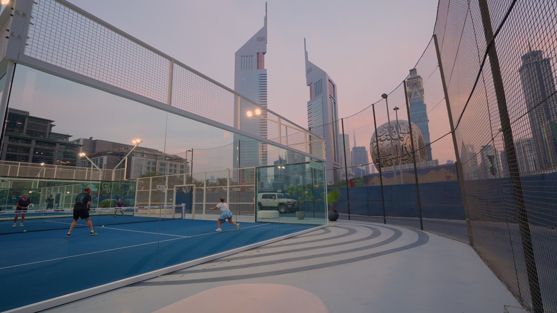 An outdoor padel court at sunset with Dubai towers behind the glass walls